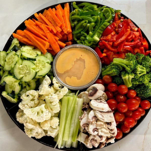 A black platter with various fresh vegetables including carrots, cucumbers, peppers, broccoli, and a dip.