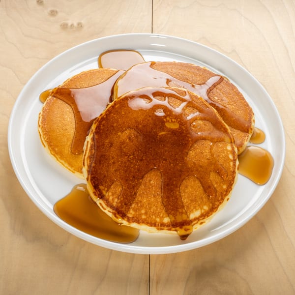 Golden pancakes drizzled with maple syrup on a white plate, on a light wooden table.