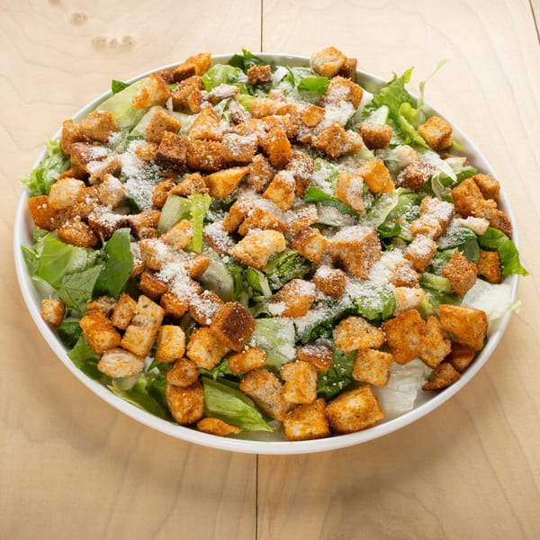 A white bowl of fresh green Caesar salad with croutons and grated cheese on a wooden table.